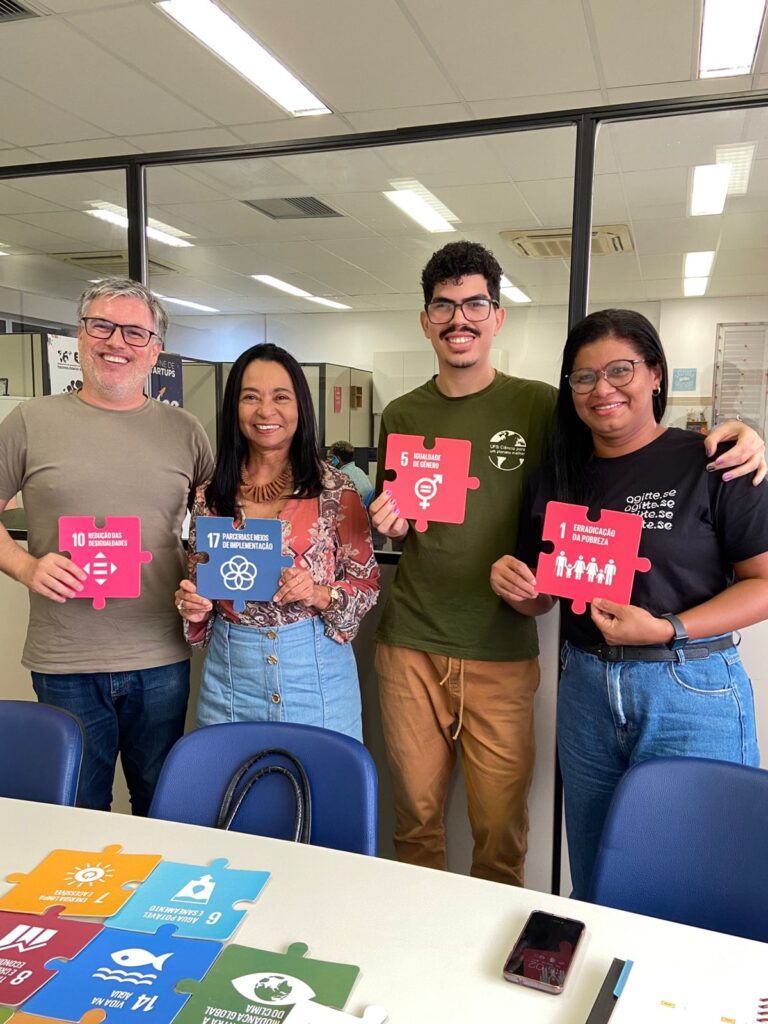 Quatro pessoas sorriem enquanto seguram peças de quebra-cabeça representando diferentes Objetivos de Desenvolvimento Sustentável (ODS), como igualdade de gênero, erradicação da pobreza, redução das desigualdades e parcerias. Elas estão em um ambiente da universidade, com mais peças dos ODS dispostas sobre a mesa à frente."