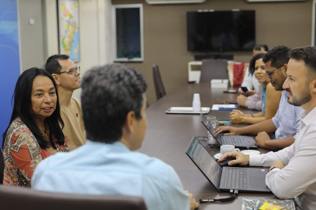 Grupo de pessoas reunidas em torno de uma mesa de reuniões, com notebooks abertos, participando de uma conversa sobre os Objetivos de Desenvolvimento Sustentável. O encontro ocorre em ambiente institucional, com destaque para a interação entre os participantes.