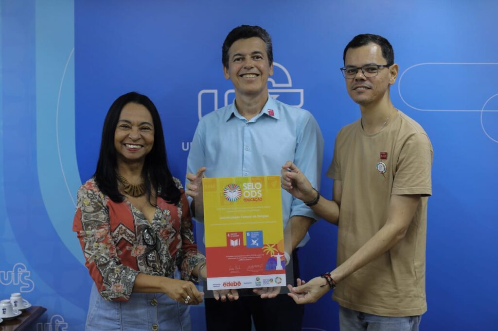 Três pessoas sorridentes posam segurando um certificado do Selo ODS Educação, com destaque para os Objetivos de Desenvolvimento Sustentável 4 (Educação de Qualidade) e 16 (Paz, Justiça e Instituições Eficazes), em frente a um painel azul com o logotipo da Universidade Federal de Sergipe.